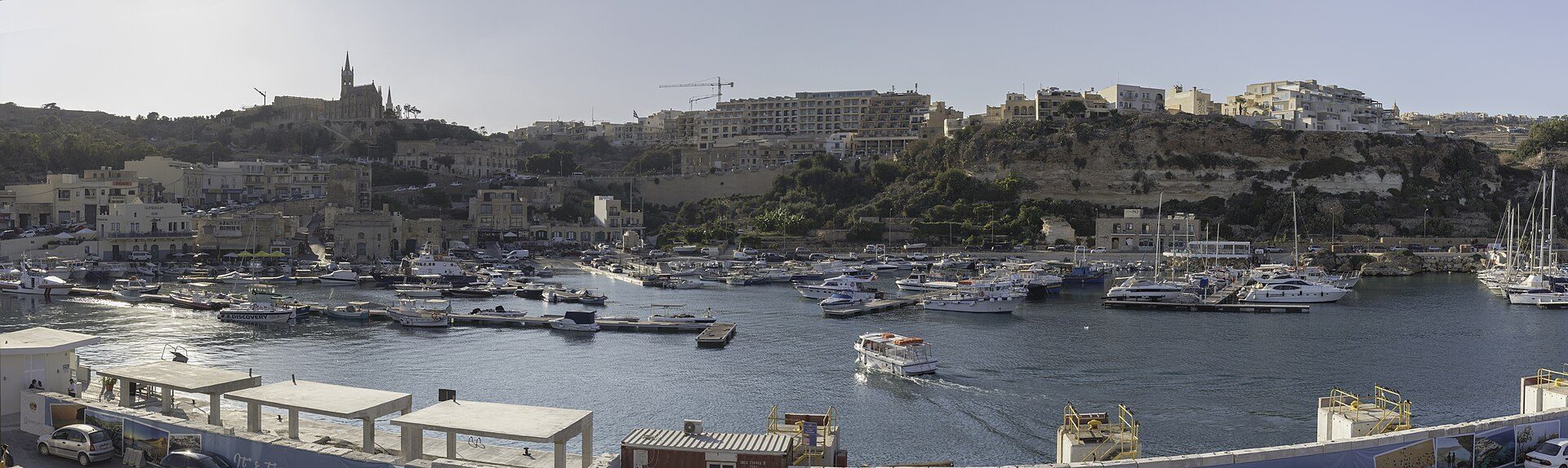 Panorama of Mġarr harbour on Gozo, boats at their moorings below the terraced hillside and the church of Our Lady of Lourdes above.