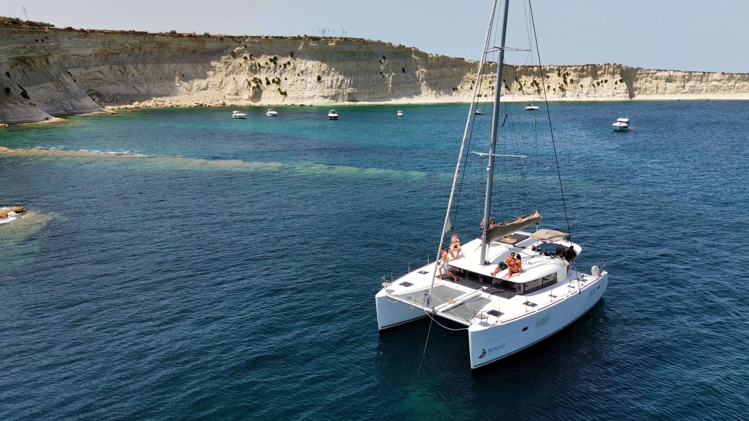 A Lagoon catamaran at anchor in clear turquoise water below Malta's limestone cliffs, guests sitting out on her foredeck.